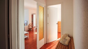 Un pasillo con un banco de mimbre, una puerta con panel texturizado y una vista parcial de una habitación con cama y cómoda.