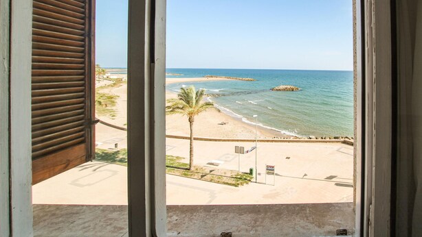 Vista a la playa desde una ventana, con vista al mar, una playa de arena y una palmera.