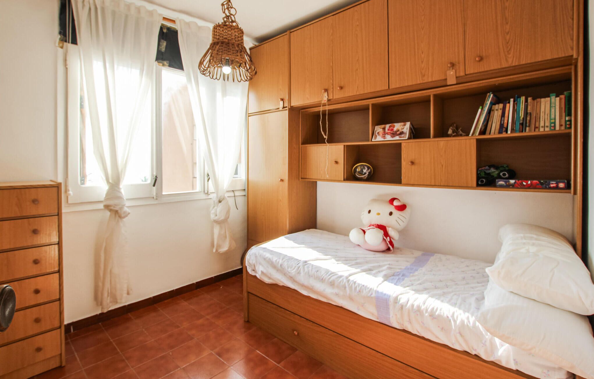 A bedroom with a wooden bed, a dresser, a window with curtains, and a shelf with books and a Hello Kitty plush toy.