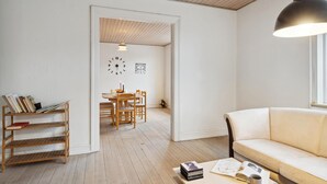 A living room with a sofa, a coffee table with books and a mug, a bookshelf, a dining area with a table and chairs, and a clock on the wall.