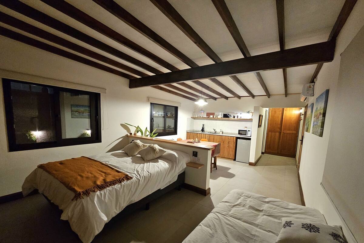 Une chambre avec un lit, une table de chevet et une fenêtre. Au fond de la pièce, on aperçoit une petite cuisine équipée d’un micro-ondes, d’un évier et de placards.