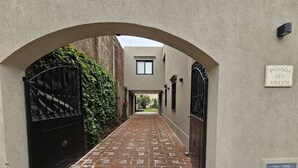A narrow alleyway with brick flooring, flanked by walls and gates, leading to a building with a sign reading "POSADA DEL ATELIER". - Posada del Atelier (San Antonio de Areco)