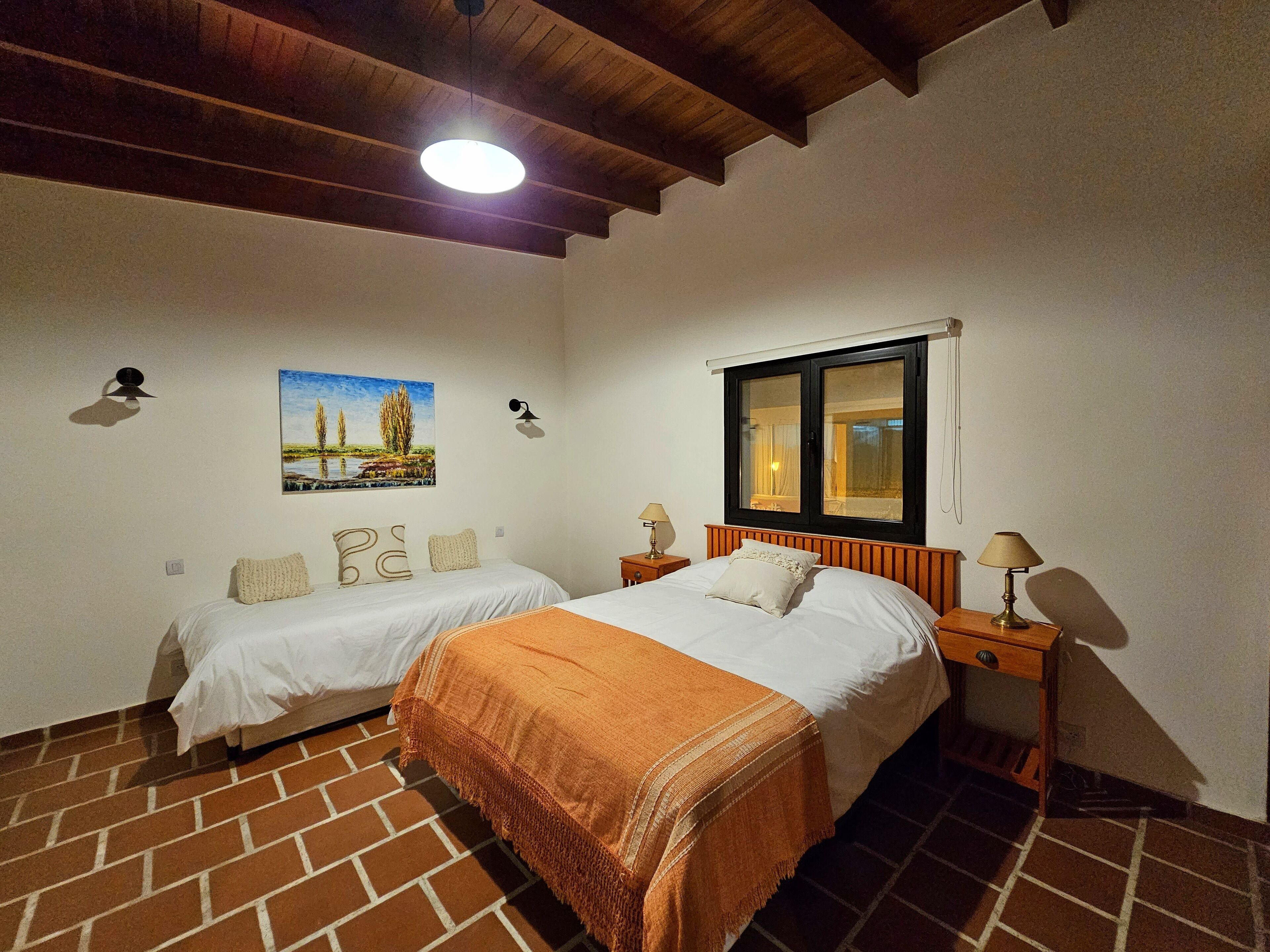A bedroom with two beds, a wooden ceiling, a window, and a painting on the wall.