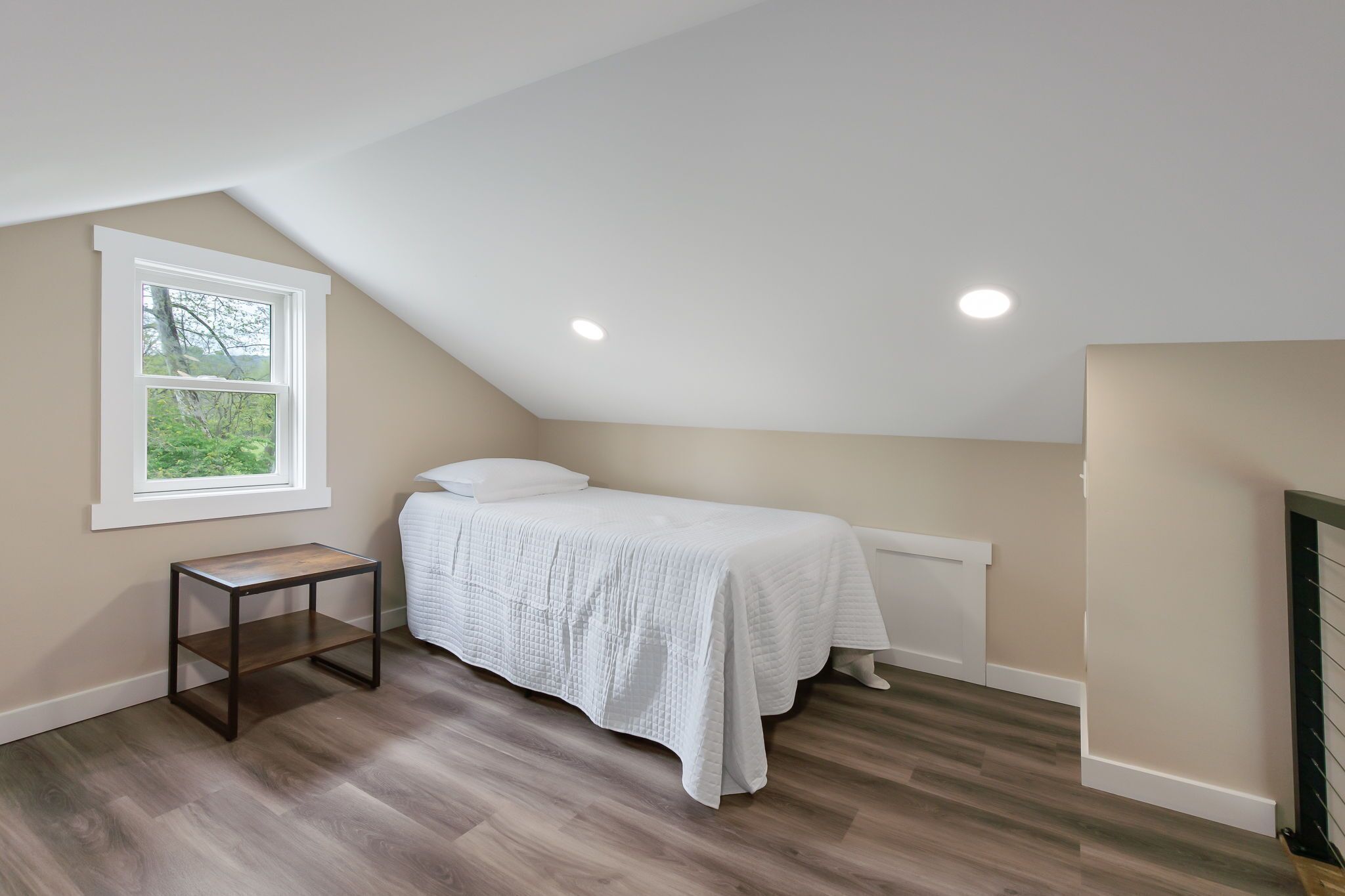 Ein Schlafzimmer mit schrägen Wänden, einem Fenster, einem Nachttisch und einem Bett mit weißer Bettdecke.