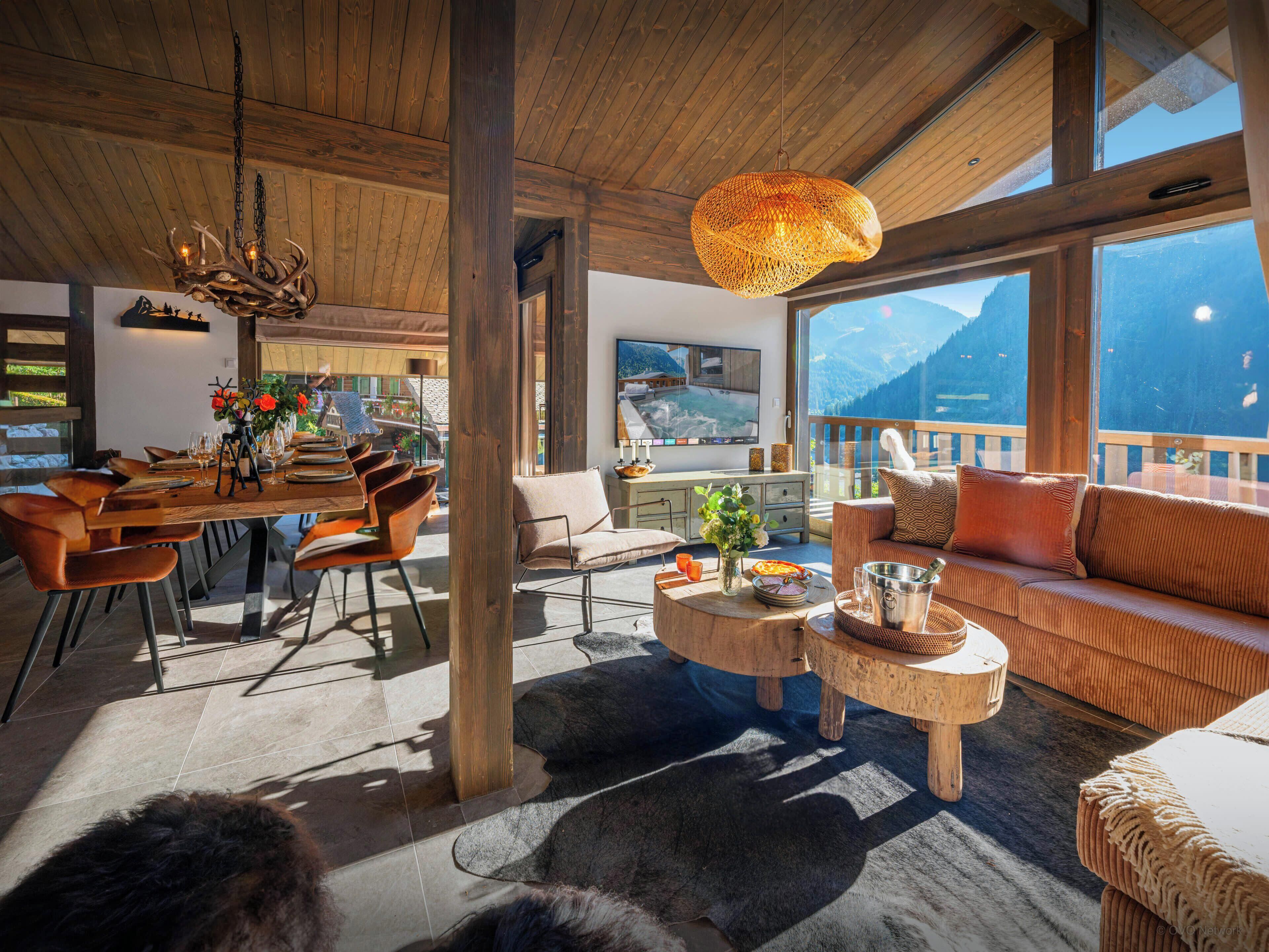 Ein geräumiges Wohnzimmer mit Holzboden, einem großen Fenster mit Blick auf die Berge, einem Essbereich mit Holztisch und -stühlen sowie einer gemütlichen Sitzecke mit Sofa und Couchtisch.
