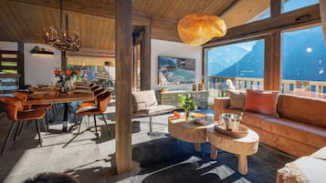 Ein geräumiges Wohnzimmer mit Holzboden, einem großen Fenster mit Blick auf die Berge, einem Essbereich mit Holztisch und -stühlen sowie einer gemütlichen Sitzecke mit Sofa und Couchtisch.