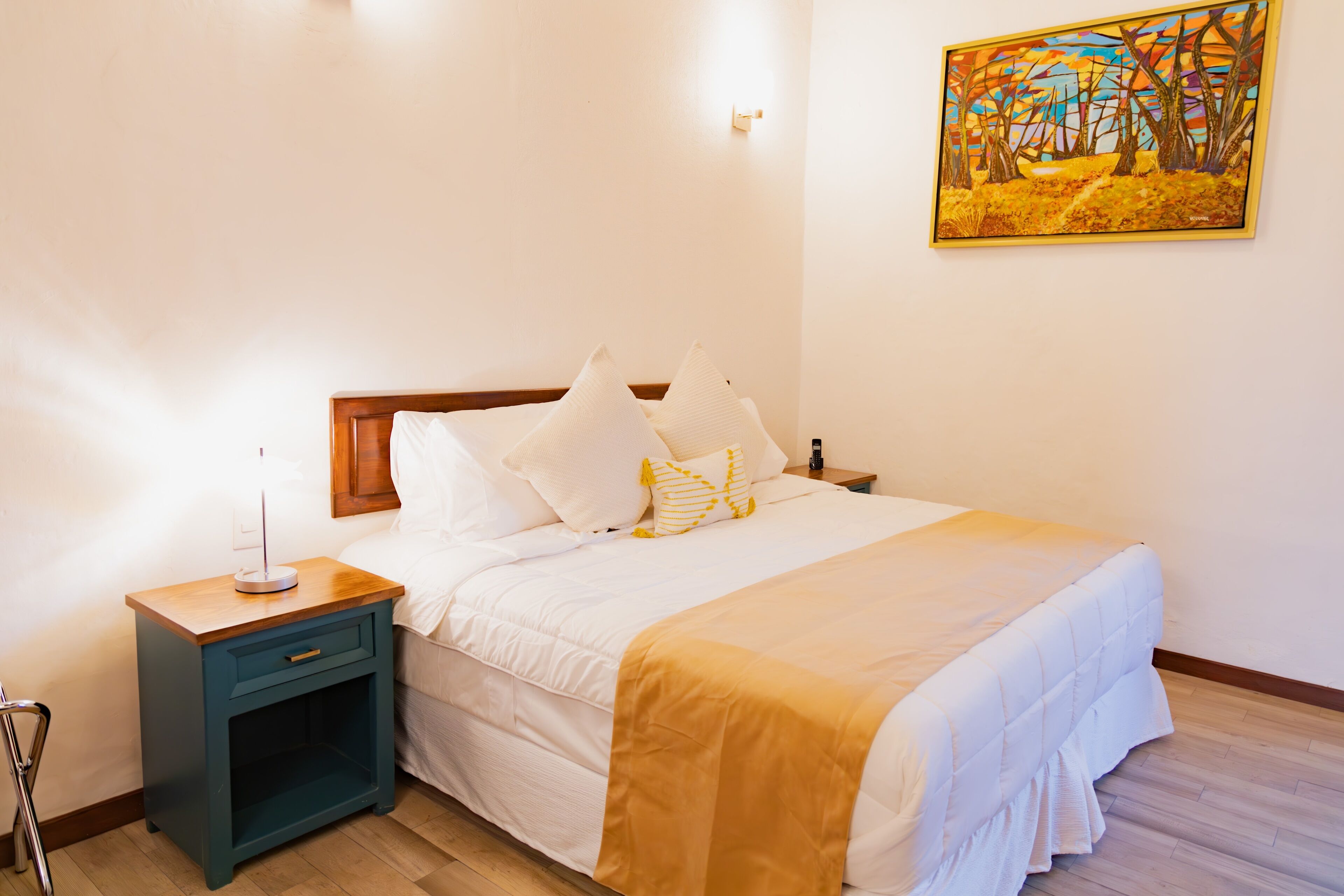 Un lit bien fait, avec du linge de lit blanc et un plaid jaune, une table de chevet avec une lampe, et un tableau accroché au mur.