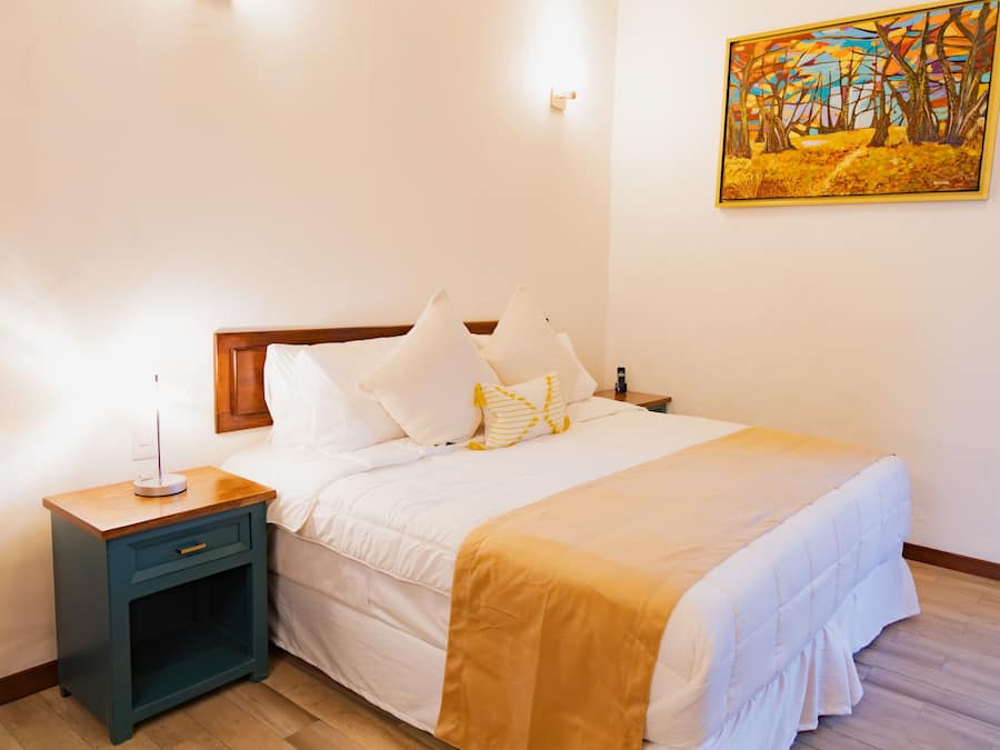 Una cama bien hecha con ropa blanca y un plaid amarillo, una mesita de noche con una lámpara y un cuadro en la pared.