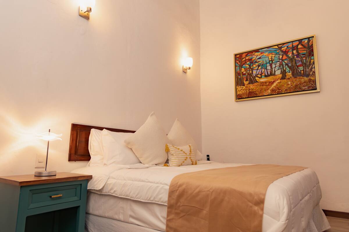A neatly made bed with white bedding and a tan throw, a teal bedside table with a lamp, and a painting on the wall.