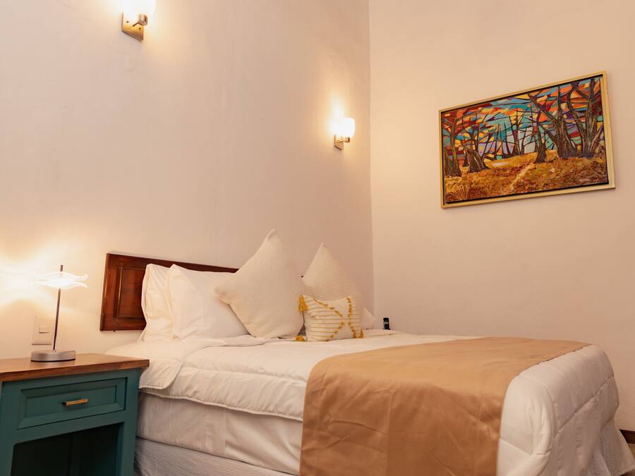 A neatly made bed with white bedding and a tan throw, a teal bedside table with a lamp, and a painting on the wall.