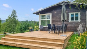 Una terraza de madera con mobiliario exterior, un porche cubierto y vistas a un jardín con flores.