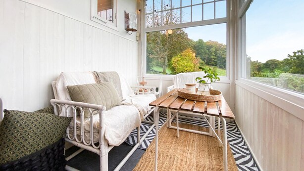 Una habitación acogedora con una cama de metal blanco, una mesa de madera con una cesta tejida, y vistas a la vegetación a través de amplios ventanales.