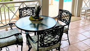 Una veranda riparata con un tavolo rotondo e quattro sedie, una ciotola di frutta sul tavolo, e vista su giardino e piscina attraverso porte aperte.