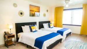 Habitación de hotel con dos camas, ventilador de techo, un cuadro y una ventana con cortinas amarillas.