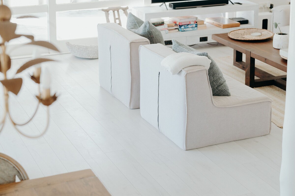 A modern living room with a flat-screen TV, white sofas, a wooden coffee table, and a large window offering a view of the outdoors.