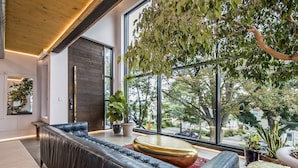 Een moderne woonkamer met een leren bank, een ronde salontafel en een groot raam met uitzicht op bomen en gebouwen.