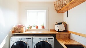 A laundry room with two front-loading washing machines, a wooden countertop, a window with potted plants, and a woven basket on a shelf. - E+H House-Views! Fast Wifi & Pet friendly! (Lyle)
