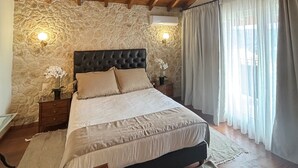 Ein Schlafzimmer mit Steinwand, Holzbalkendecke, einem Bett mit Kopfteil, Nachttischen mit Lampen und einem Fenster mit Vorhängen.