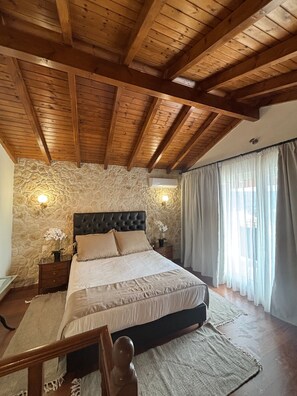 A bedroom with a stone wall, wooden ceiling beams, a bed with a headboard, bedside tables with lamps, and a window with curtains.
