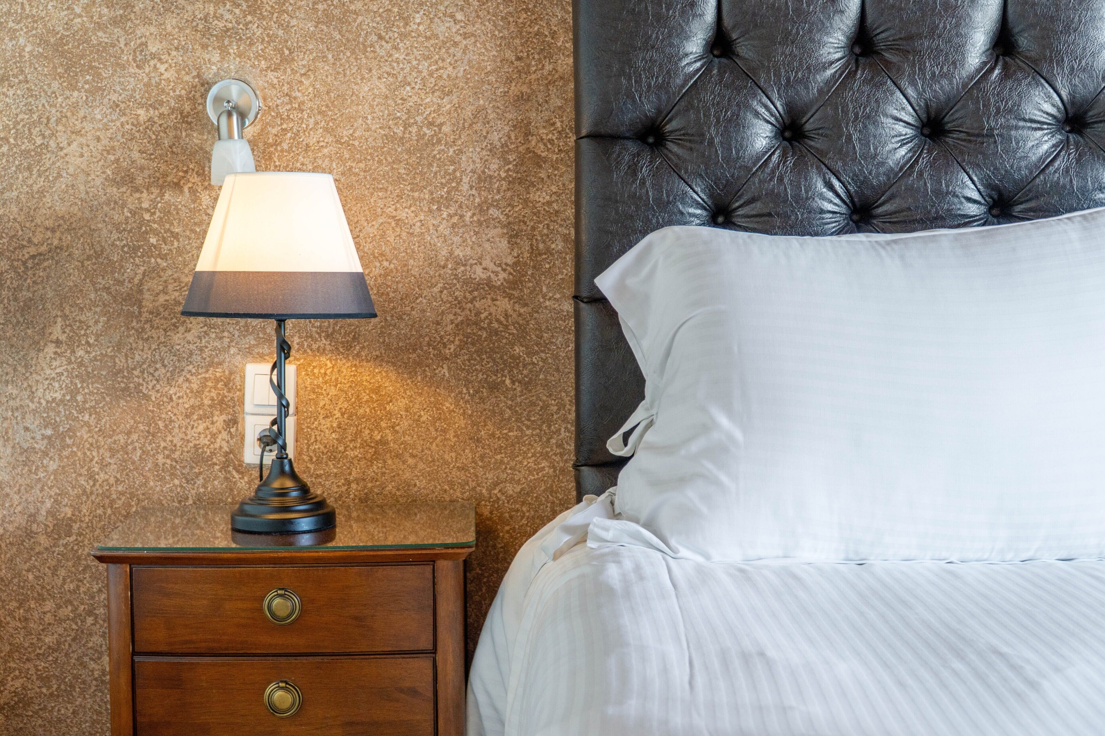 Una cama con sábanas blancas, un cabecero capitonado negro, una mesita de noche de madera con una lámpara y una pared con textura.