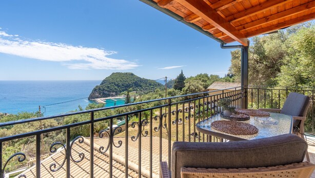 Ein Balkon mit Glastisch und Stühlen, der einen malerischen Blick auf das Meer und eine gebirgige Insel bietet.