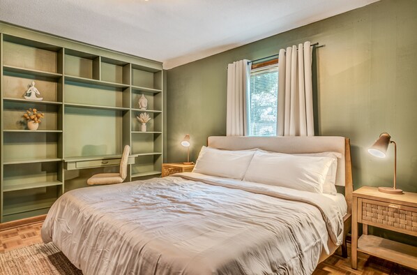 A bedroom with a large bed, a built-in bookshelf, a nightstand with a lamp, and a window with curtains.