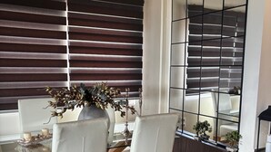 Un comedor con una mesa de vidrio, sillas blancas, un jarrón con flores secas y una ventana con persianas oscuras.