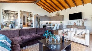 Un salón con un sofá marrón oscuro, una mesa de centro con objetos decorativos, un sillón blanco con cojines de estampado de leopardo y un techo de madera.