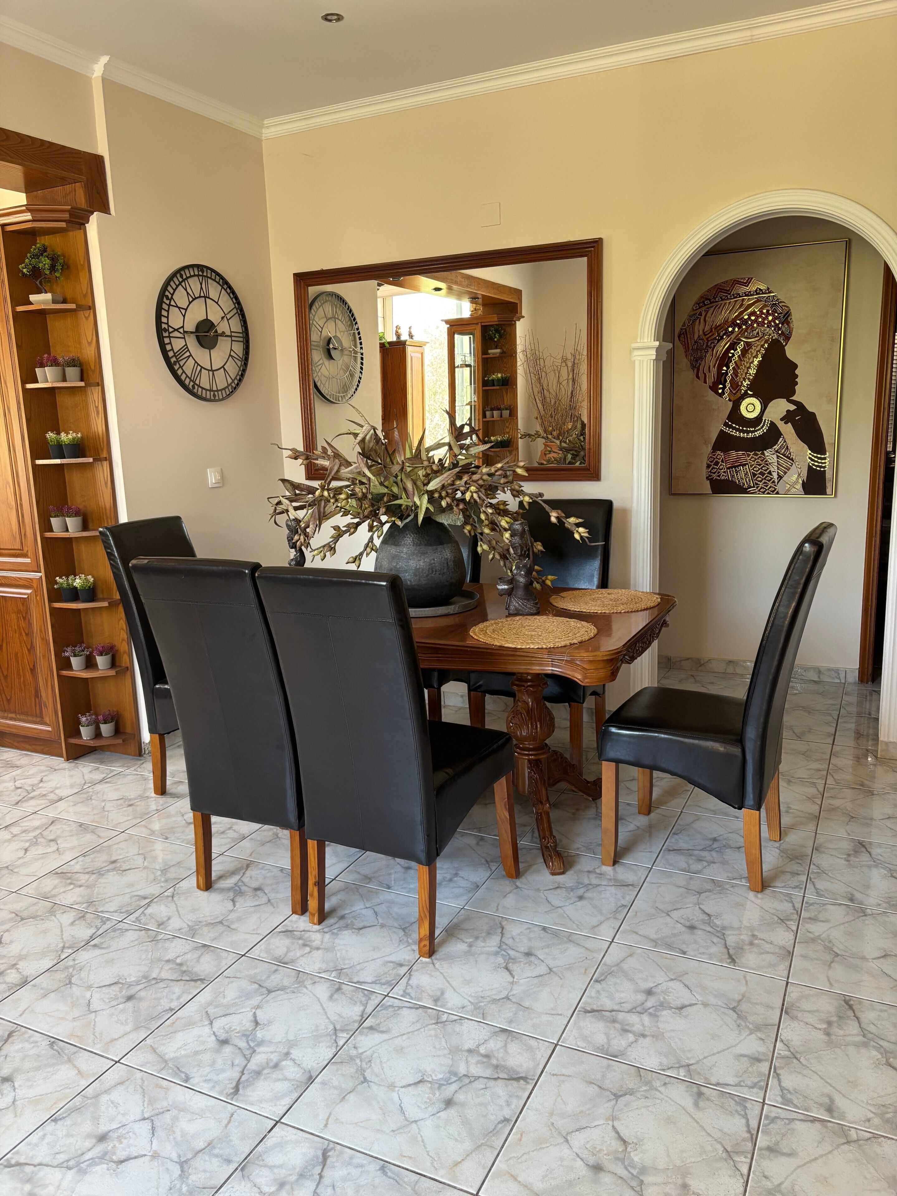 A dining area with a wooden table, black chairs, a vase with plants, a clock, and a framed artwork.