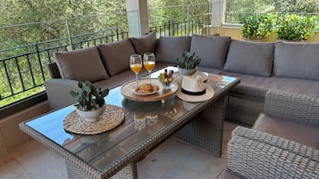 A sunlit patio with wicker furniture, a glass-top table, and a view of greenery.