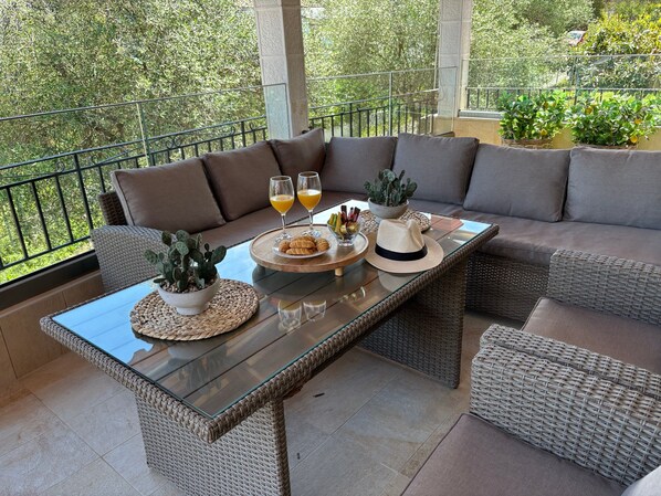 A sunlit patio with wicker furniture, a glass-top table, and a view of greenery.