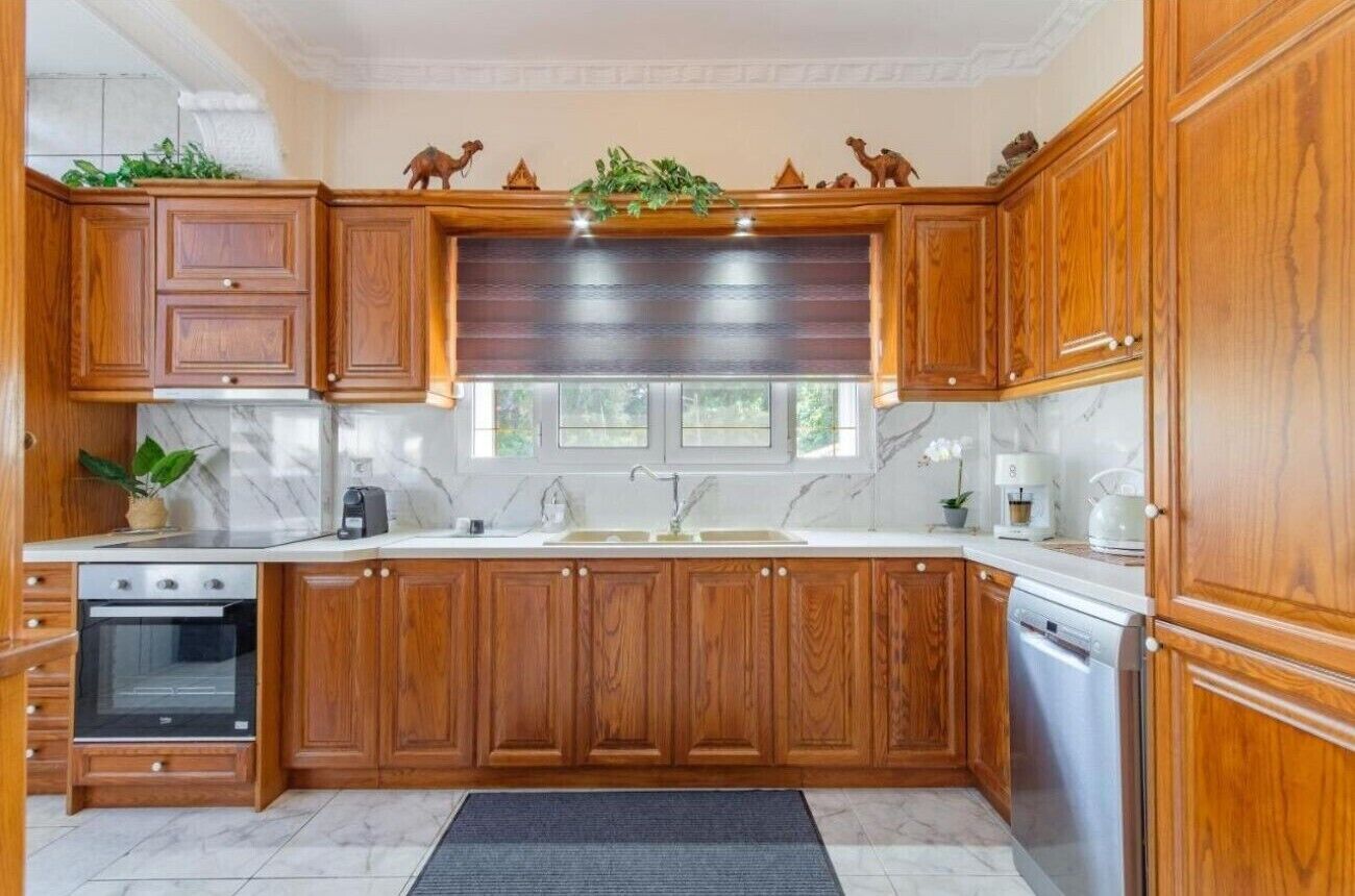 Una cocina con armarios de madera, encimera de mármol, horno empotrado, lavavajillas y ventana con vistas a la vegetación.