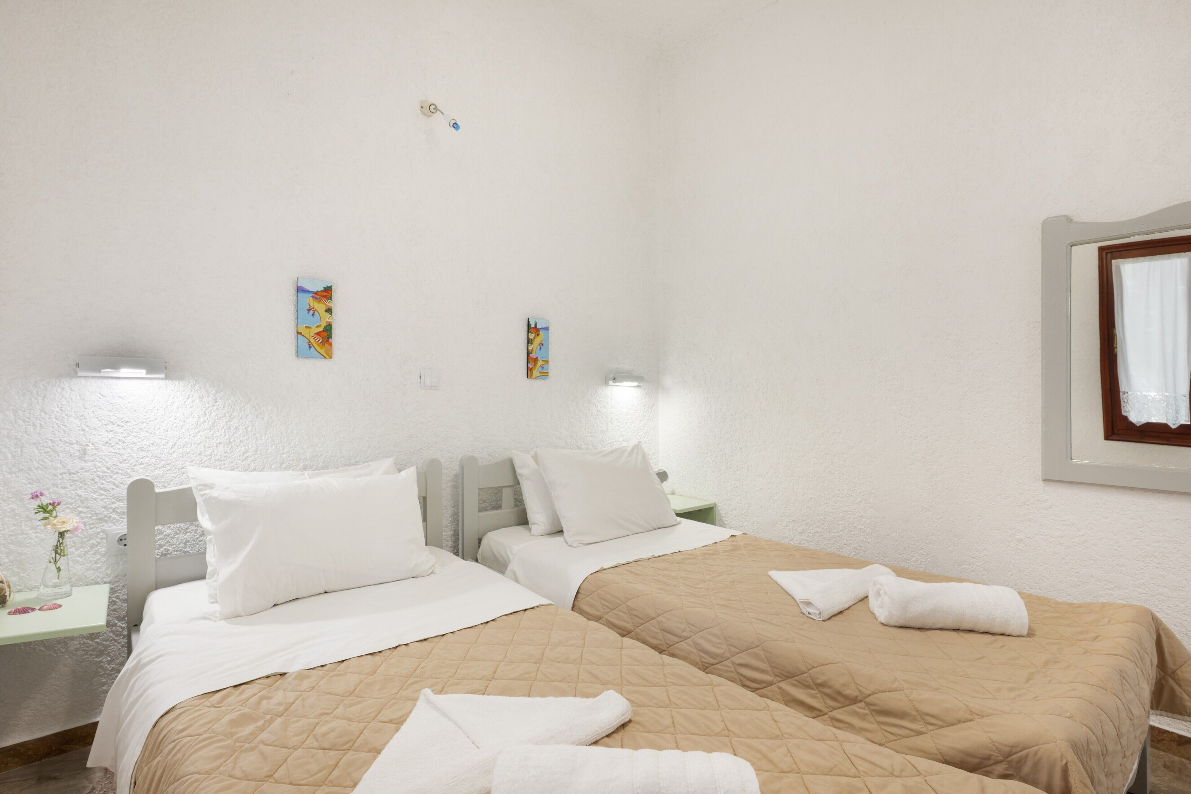 Une chambre d’hôtel avec deux lits, du linge de lit blanc, une petite table de chevet ornée d’un vase de fleurs et deux lampes fixées au mur.