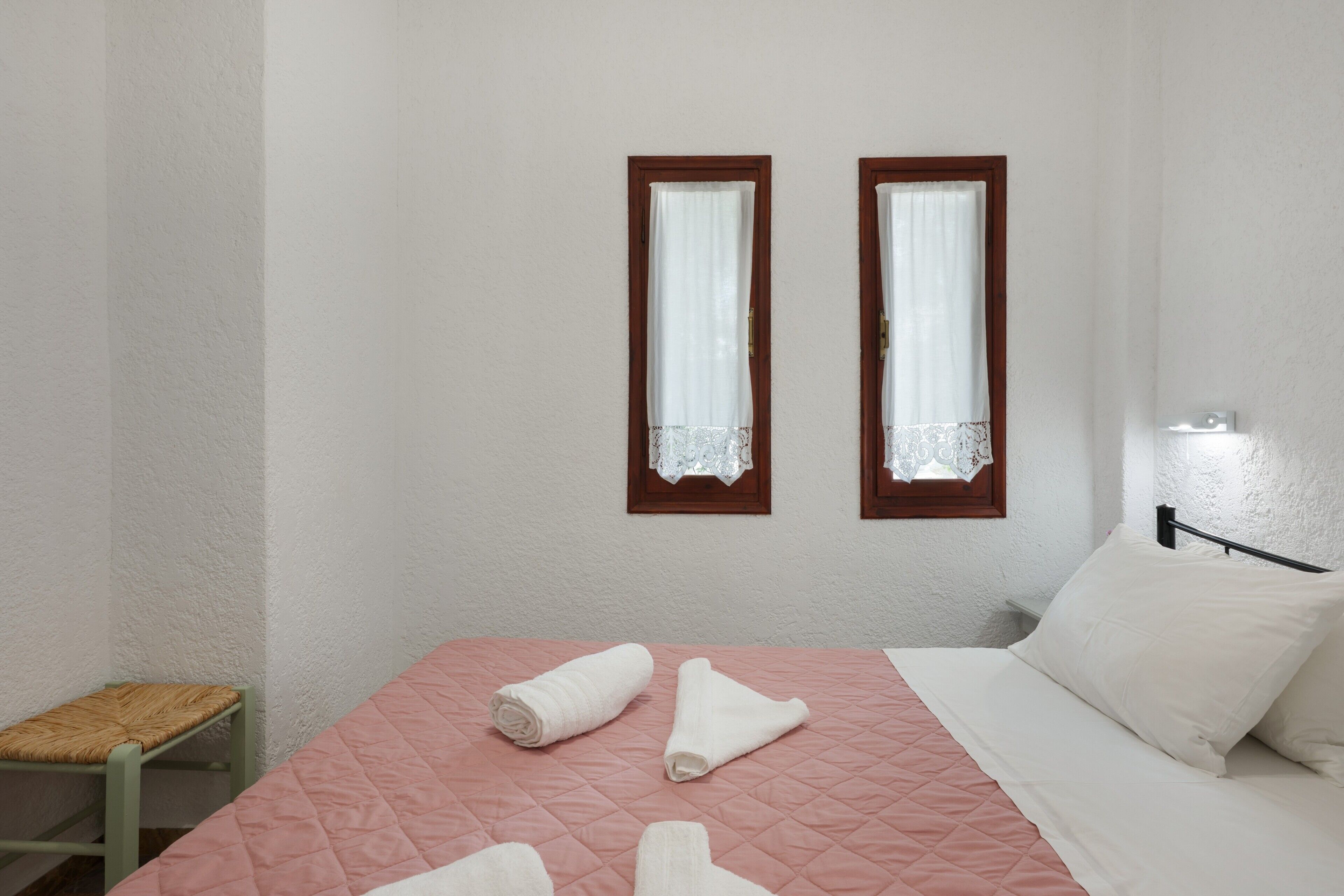 Ein Schlafzimmer mit einem Bett, zwei Fenstern mit Spitzenvorhängen, einem Nachttisch und einer Lampe.