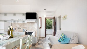 A modern kitchen with a flat-screen TV, a dining area with a table and chairs, a sofa with a patterned pillow, and a view of greenery outside.
