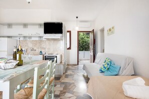 Eine moderne Küche mit Flachbildfernseher, ein Essbereich mit Tisch und Stühlen, ein Sofa mit einem gemusterten Kissen und Blick ins Grüne.