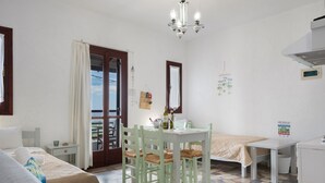 A room with a bed, a table with chairs, a small kitchenette, a window with curtains, and a chandelier.