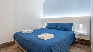 A neatly made bed with blue bedding and white towels, a bedside table with a light, and a wooden floor.