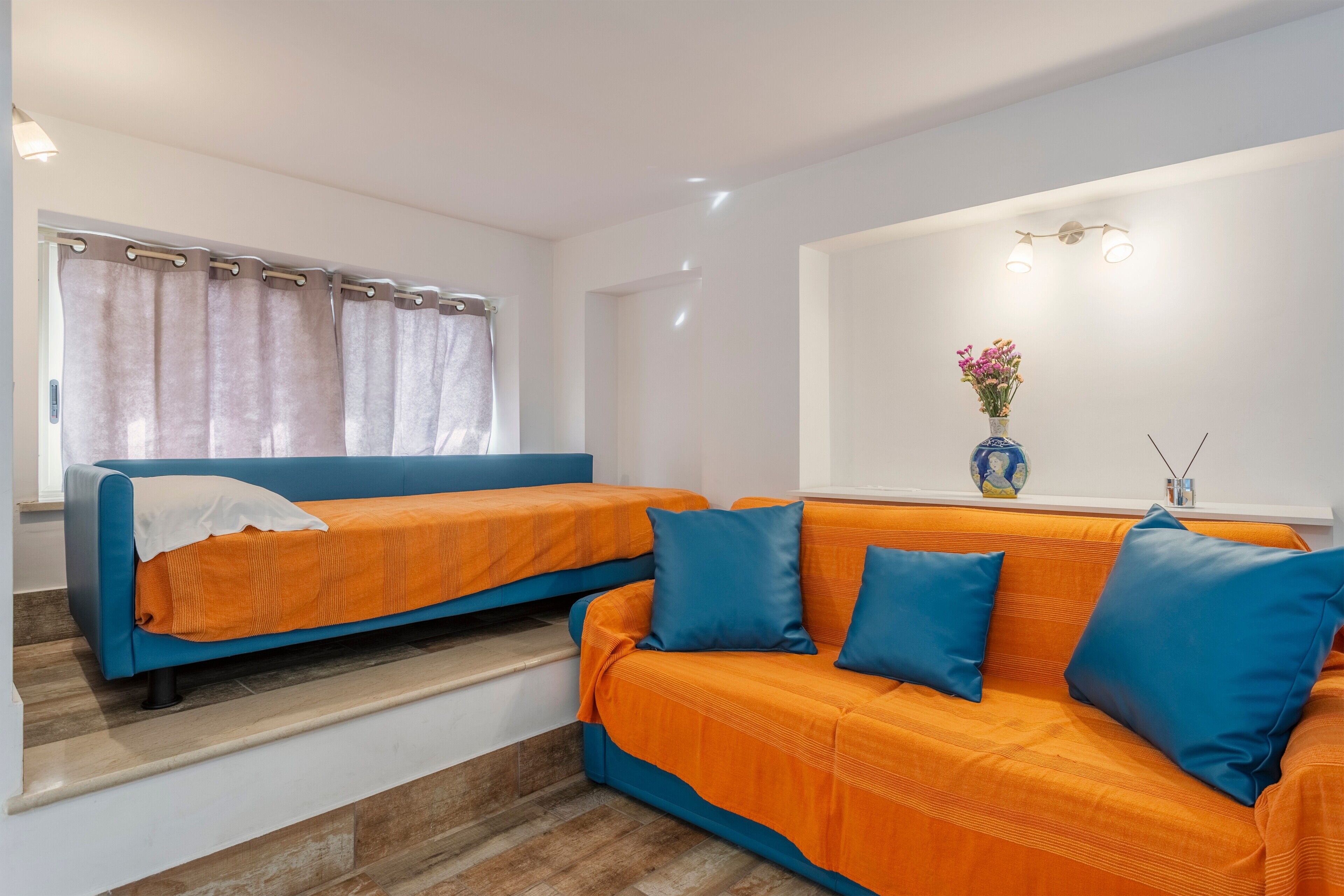 Une pièce avec deux canapés, une fenêtre avec des rideaux, un vase de fleurs et un luminaire fixé au mur.