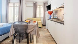Quarto de hotel compacto com cama, mesa de jantar com cadeira, pequena área de cozinha e janela com cortinas.