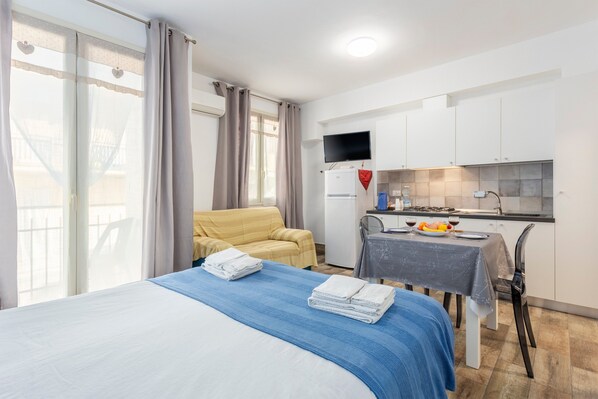 A hotel room with a bed, a sofa, a dining table with a chair, a kitchen area, and a TV mounted on the wall.
