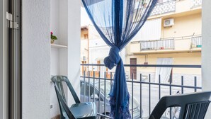 Uma varanda com cortina azul, duas cadeiras de plástico e vista para uma rua com edifícios.