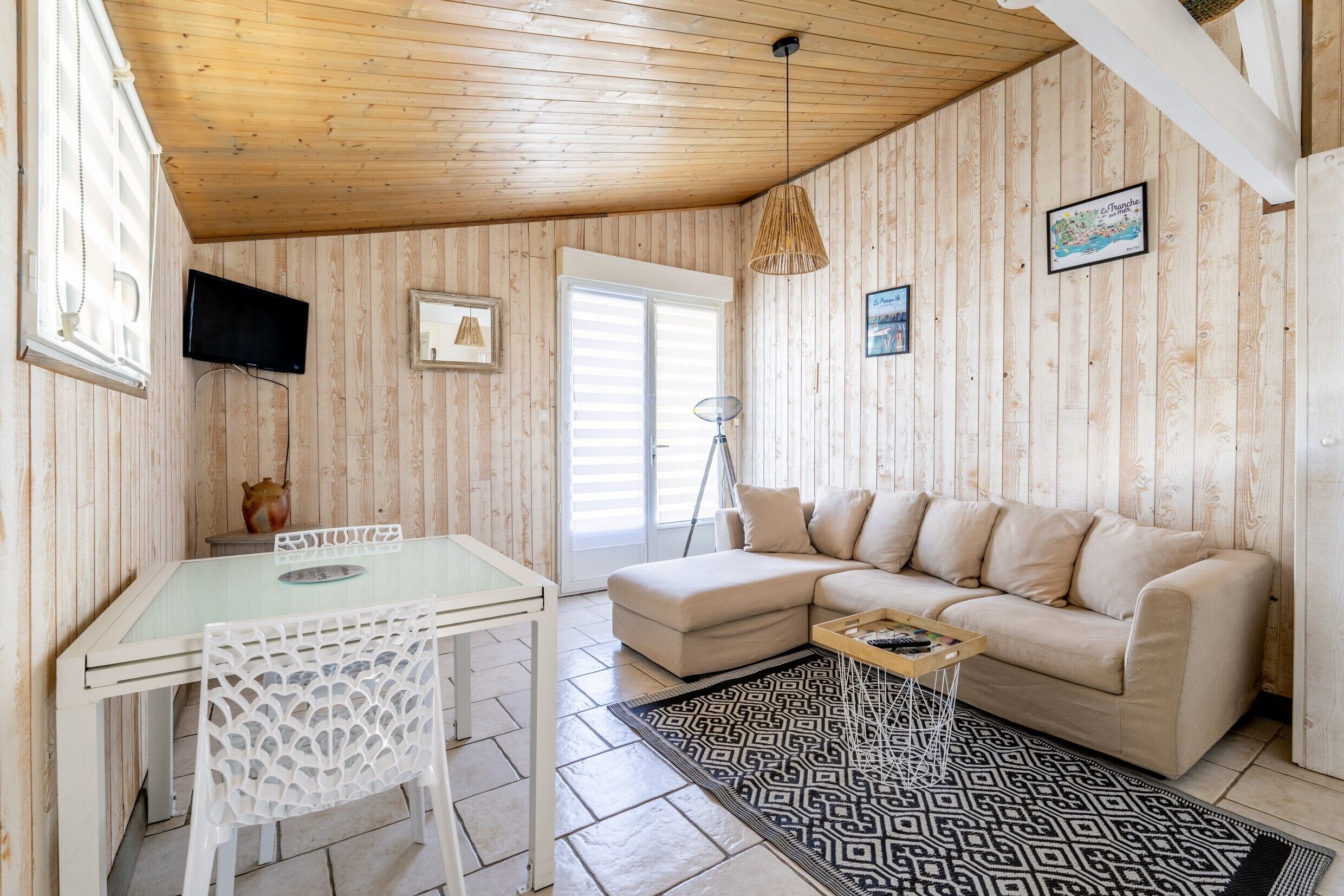 Un salón acogedor con techo de madera, una mesa de vidrio, una silla blanca, un sofá color beige y una alfombra estampada.