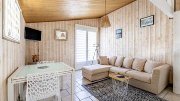 Un salón acogedor con techo de madera, una mesa de vidrio, una silla blanca, un sofá color beige y una alfombra estampada.