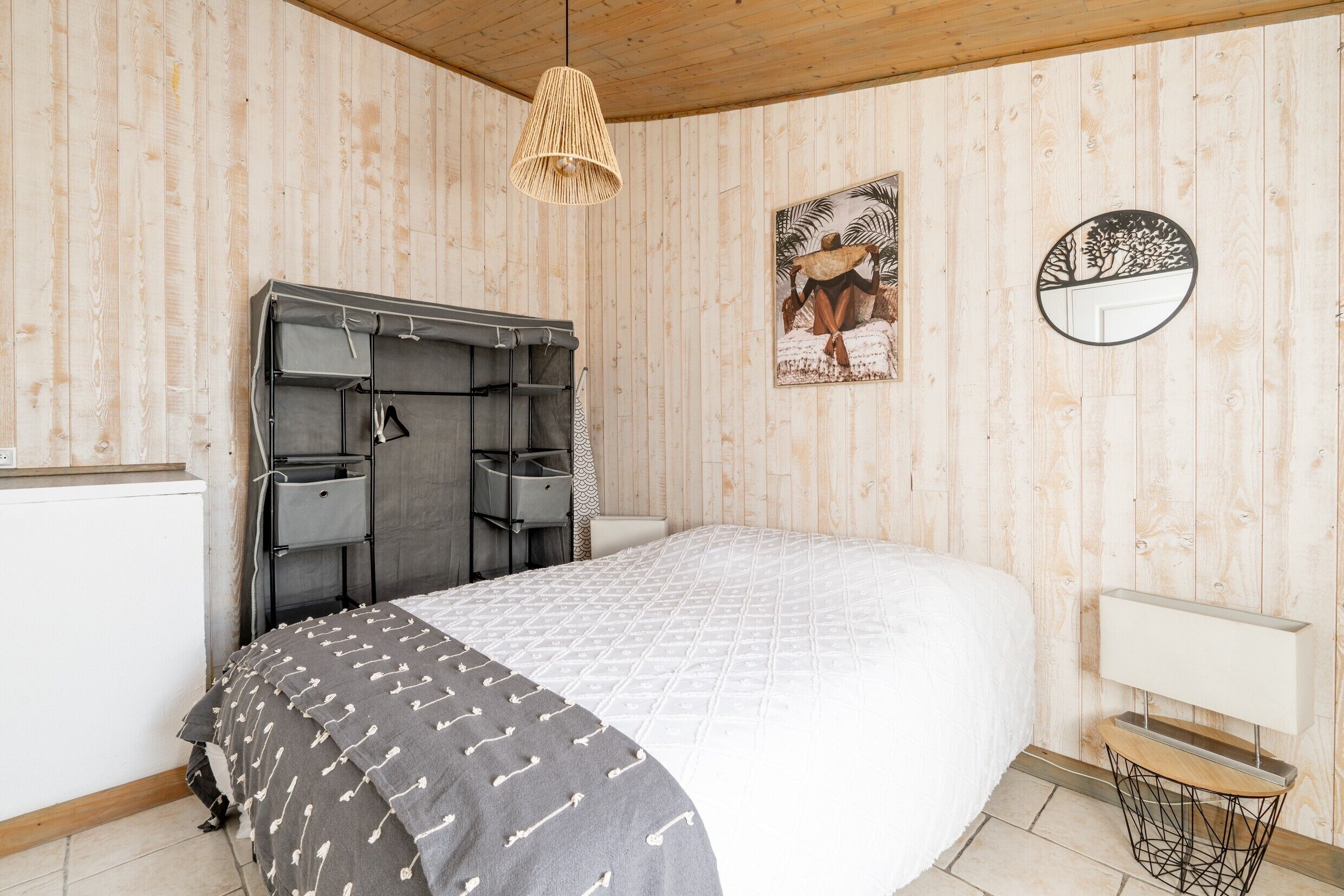 A bedroom with a bed, a hanging light fixture, a wall-mounted artwork, and a round mirror.