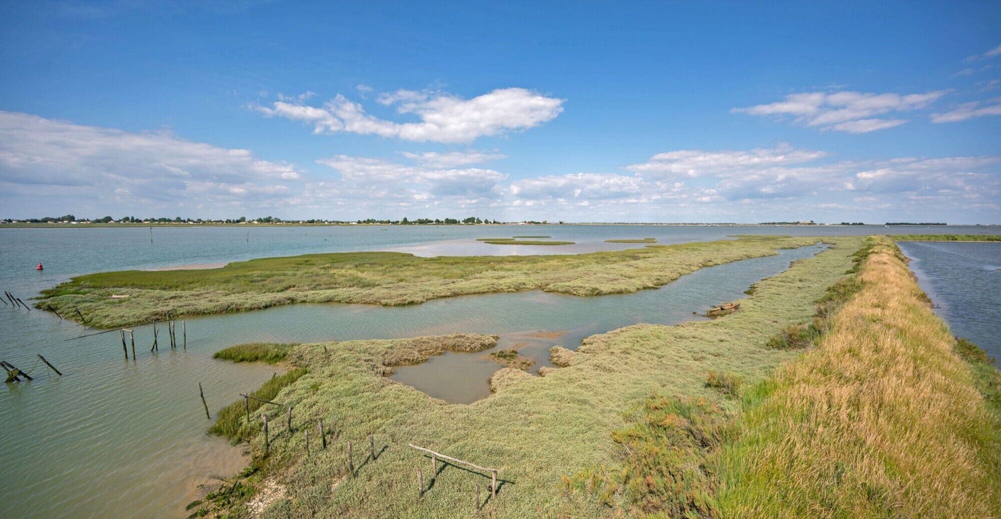 Een kustlandschap met een waterweg, grasvelden en een verre kustlijn.