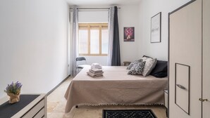 Een slaapkamer met een bed, nachtkastje, stoel, raam met gordijnen en een aan de muur hangend schilderij.