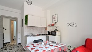Een kleine keuken met witte kastjes, een magnetron en een koffieapparaat. Er is een eettafel met een tafelkleed met een hartprint en een rode stoel.