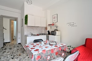 A compact kitchen with white cabinets, a microwave, and a coffee machine. There is a dining table with a heart-patterned tablecloth and a red chair. - Vacation Home "Jerzu Centro" with Private Terrace & Balcony (Jerzu)