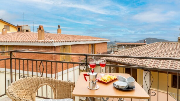 A balcony with a table set for two, a wicker chair, and a view of buildings and mountains. - Vacation Home "Jerzu Centro 2" with Private Terrace & Balcony (Jerzu)